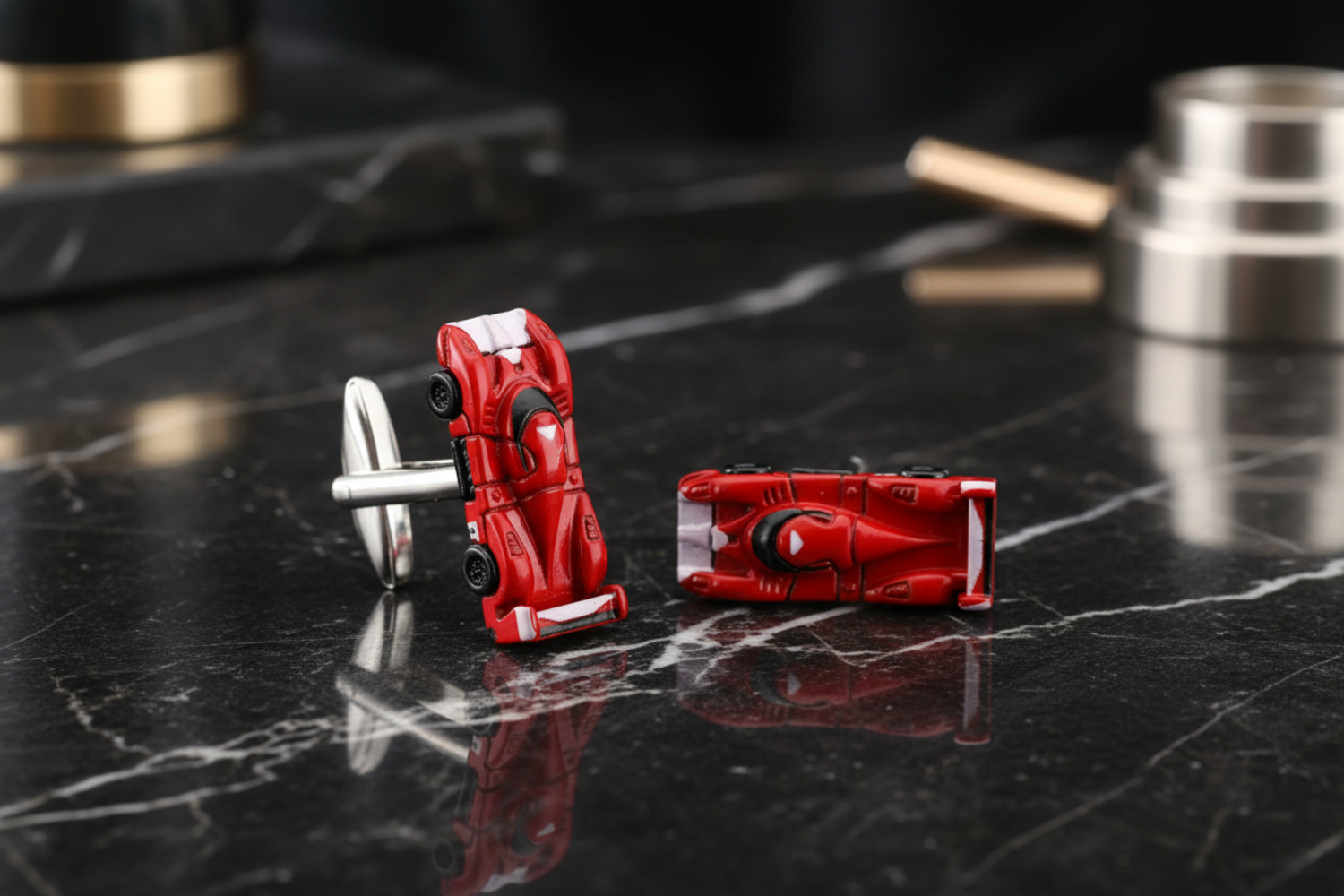 A pair of red, black, and chrome race car-shaped cufflinks placed on a wooden surface with green leafy elements in the background.