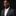 Man wearing a dark suit with a white shirt and a small flag pin on a black background