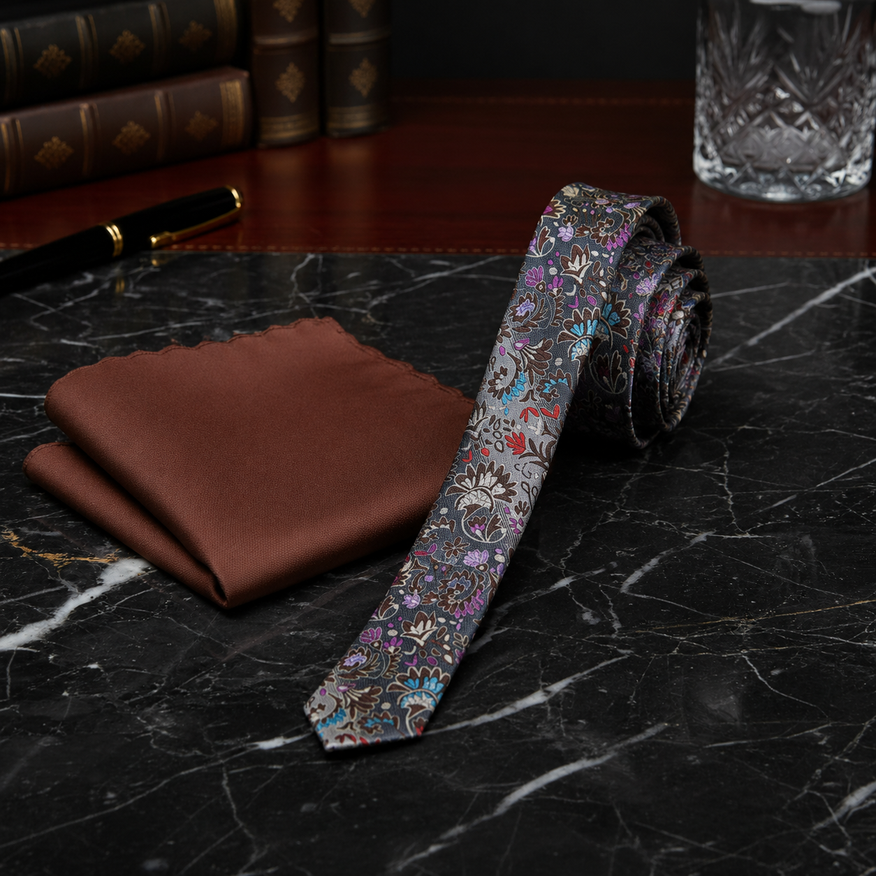 A men's necktie with an intricate multicolor paisley and floral pattern, displayed next to a matching brown silk pocket square on a black marble surface.