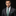 Man wearing a gray suit with a green and blue plaid tie and pocket square on a black background