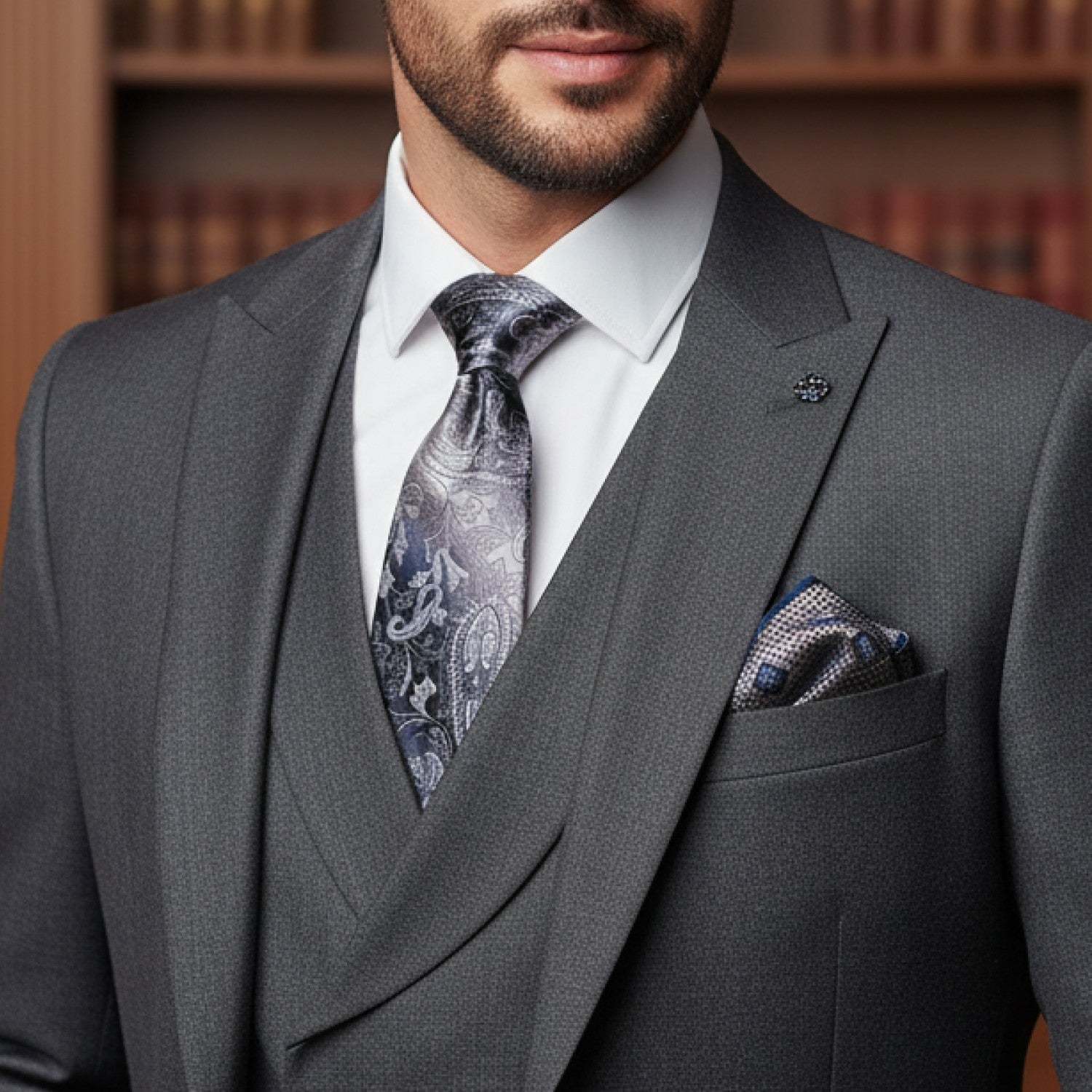 Man wearing a gray suit with a patterned tie and pocket square against a bookshelf background