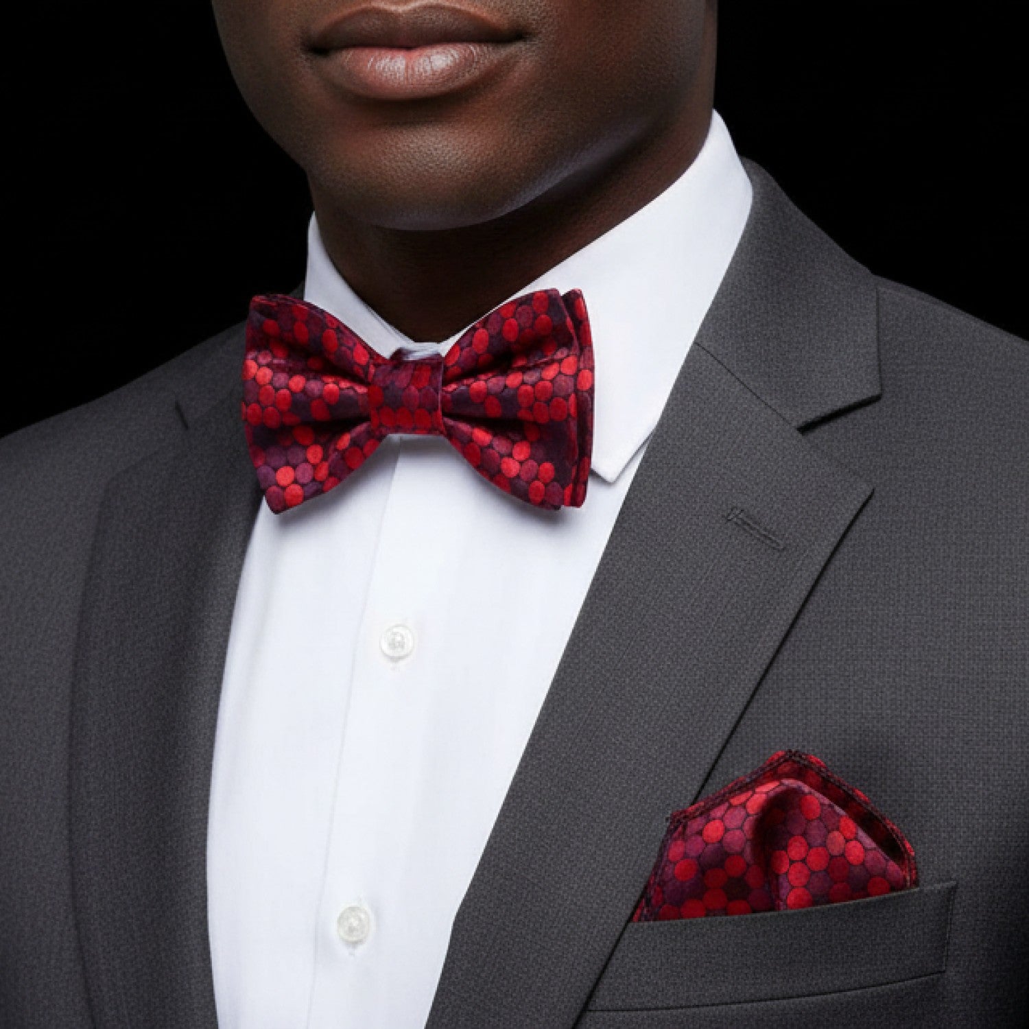 Man wearing a gray suit with a red patterned polka bow tie and pocket square on a black background