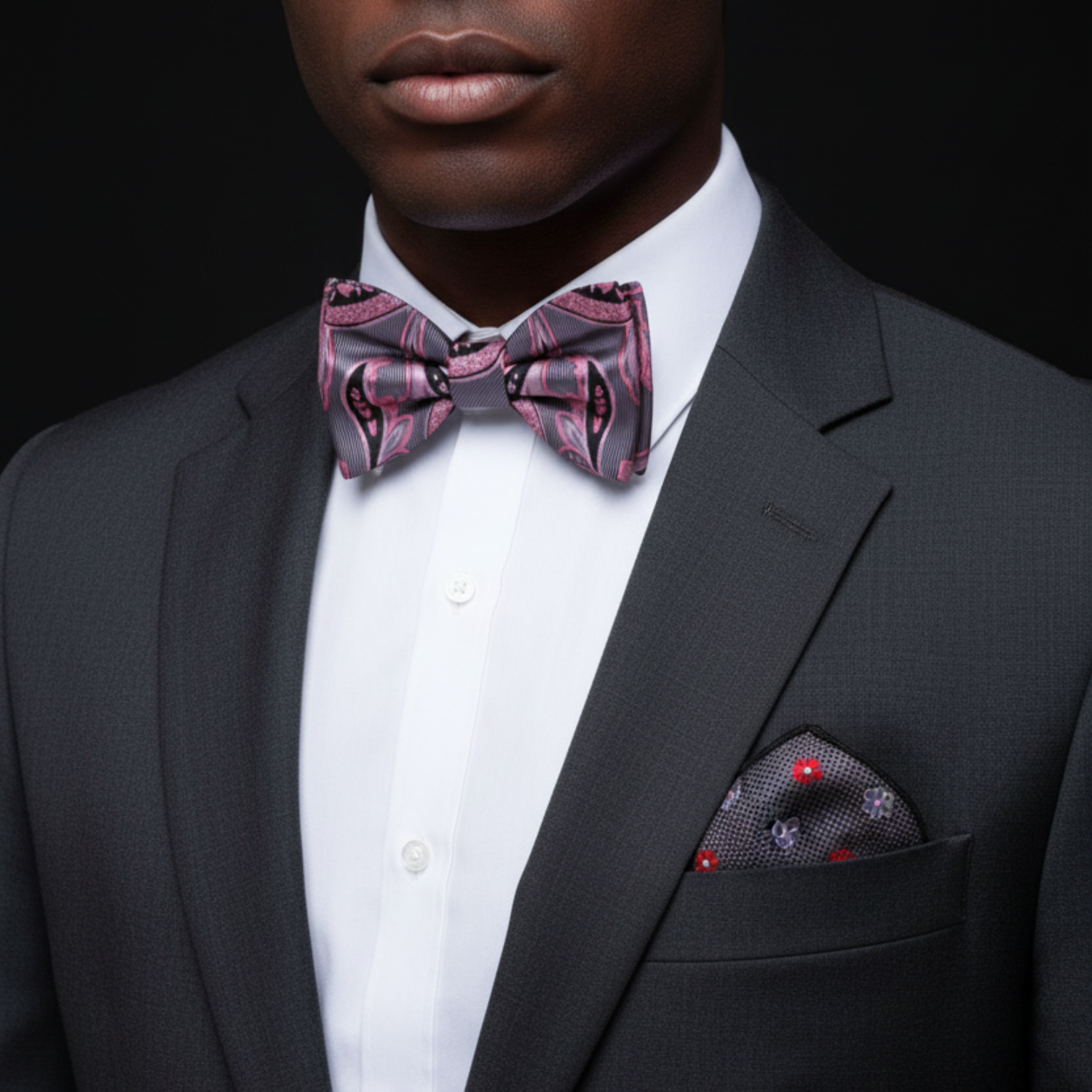 Man wearing a gray suit with a pink bow tie and pocket square against a black background