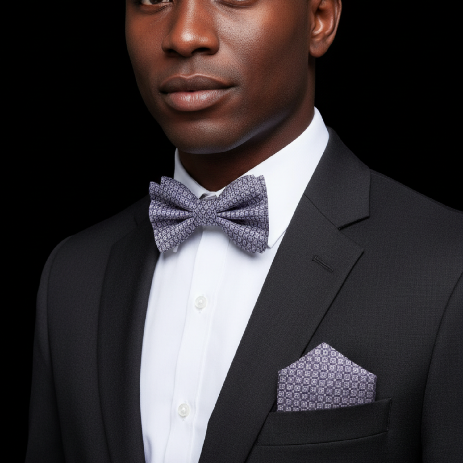 Man wearing a black suit with a white shirt and patterned bow tie and pocket square on a black background
