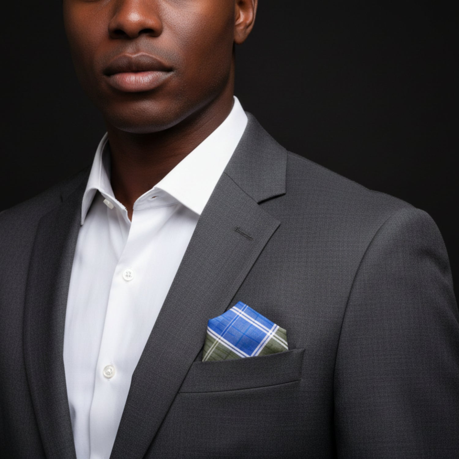 Man wearing a gray suit with a white shirt and blue plaid pocket square on a black background