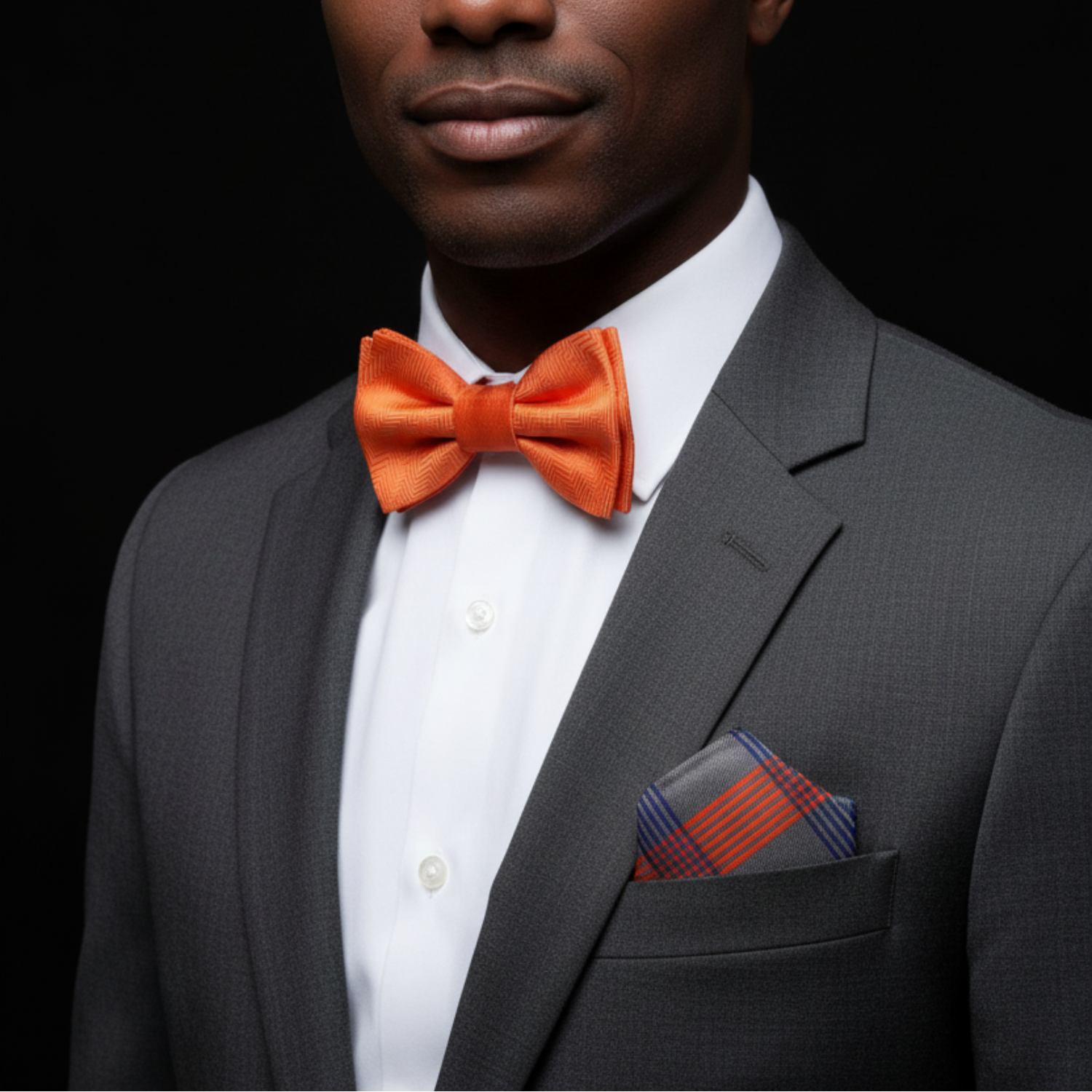Man wearing a gray suit with an orange bow tie and plaid pocket square on a black background