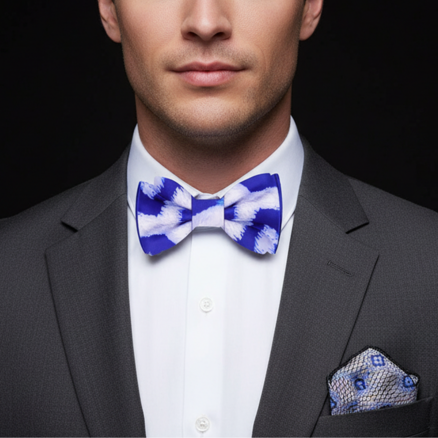 Man wearing a gray suit with a blue and white patterned bow tie and pocket square on a black background