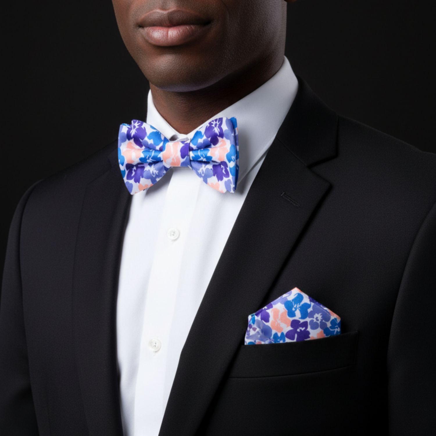 Man wearing a black suit with a colorful floral bow tie and pocket square on a black background