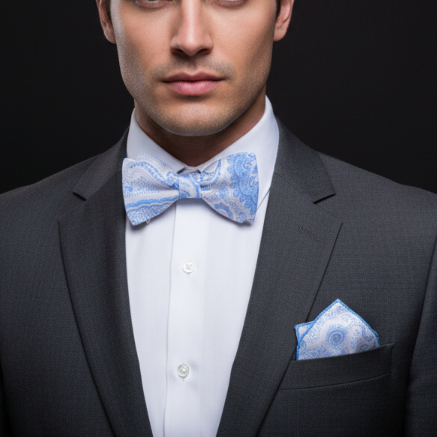 Man wearing a gray suit with a white shirt, blue patterned bow tie, and pocket square against a black background.