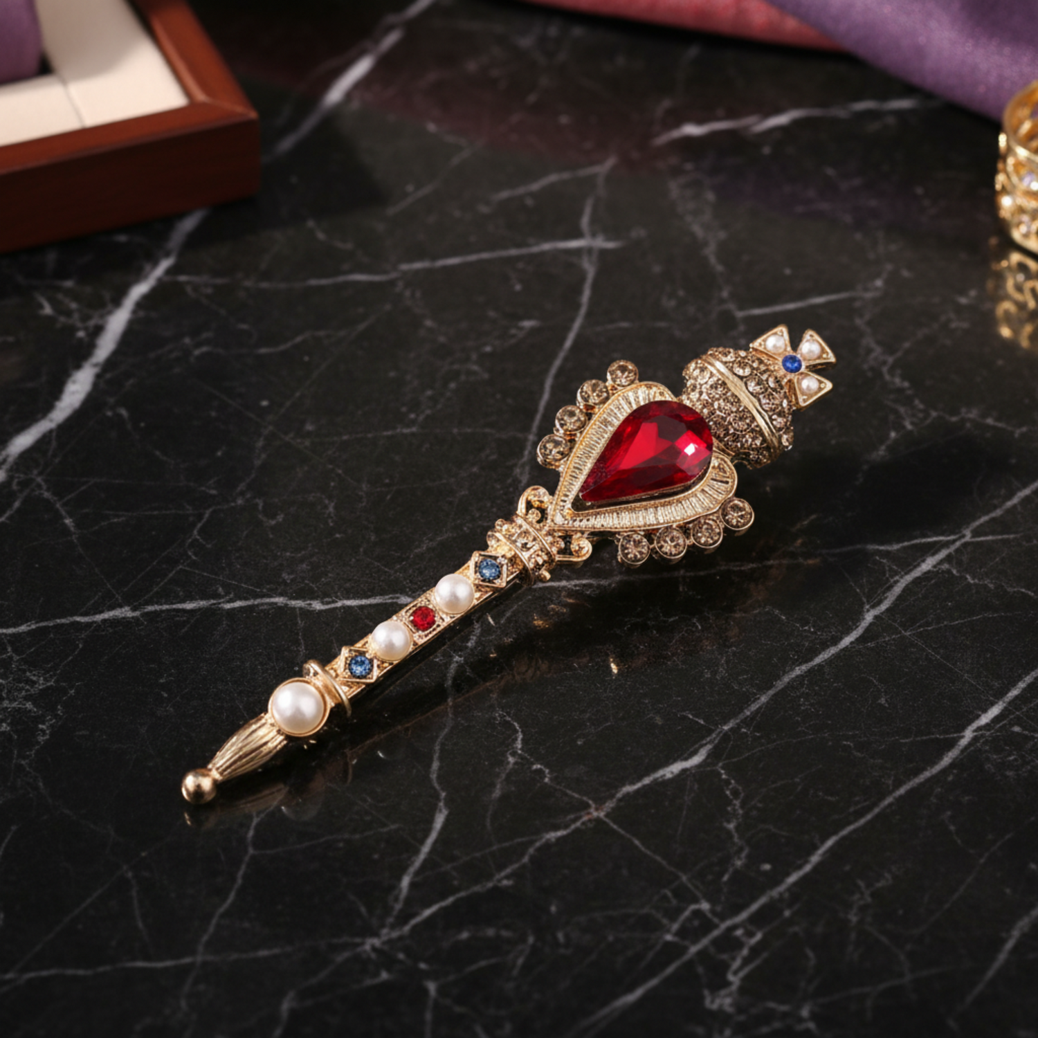 A decorative crown-shaped brooch or pin in gold-tone metal with a large red teardrop crystal, small clear stones, faux pearls, and blue and red accent crystals, shown on a white background.