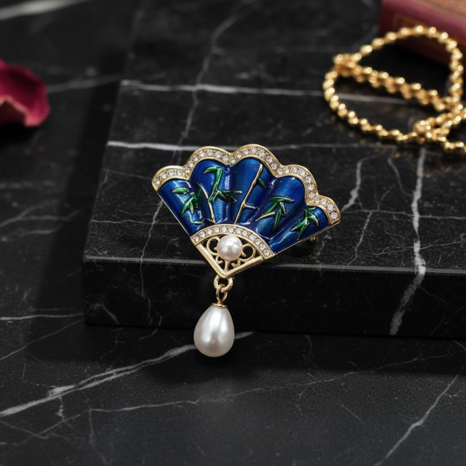 A decorative brooch shaped like a fan, featuring blue enamel with green tropical leaf motifs, gold-toned metal, clear rhinestones, and a dangling white pearl, styled on a kraft paper surface with a book and green leaves in the background.