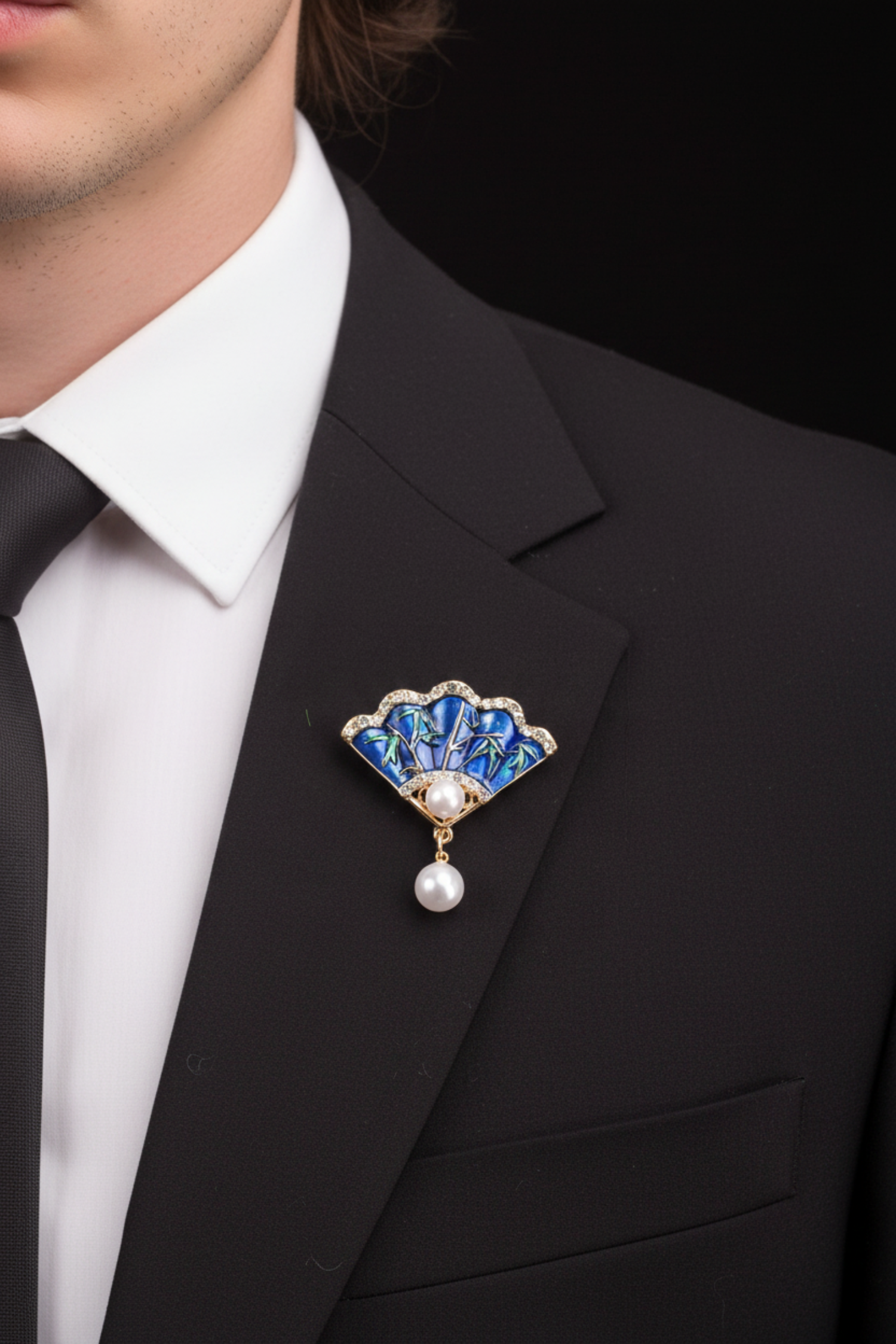 A gold-tone fan-shaped brooch with a blue enamel background, green bamboo leaf details, a central white pearl, and a dangling white pearl at the bottom, set against a black background. 