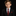 A young boy with light brown hair is wearing a black suit jacket, a white dress shirt, a textured dark red tie, and a matching dark red pocket square, set against a solid black background.
