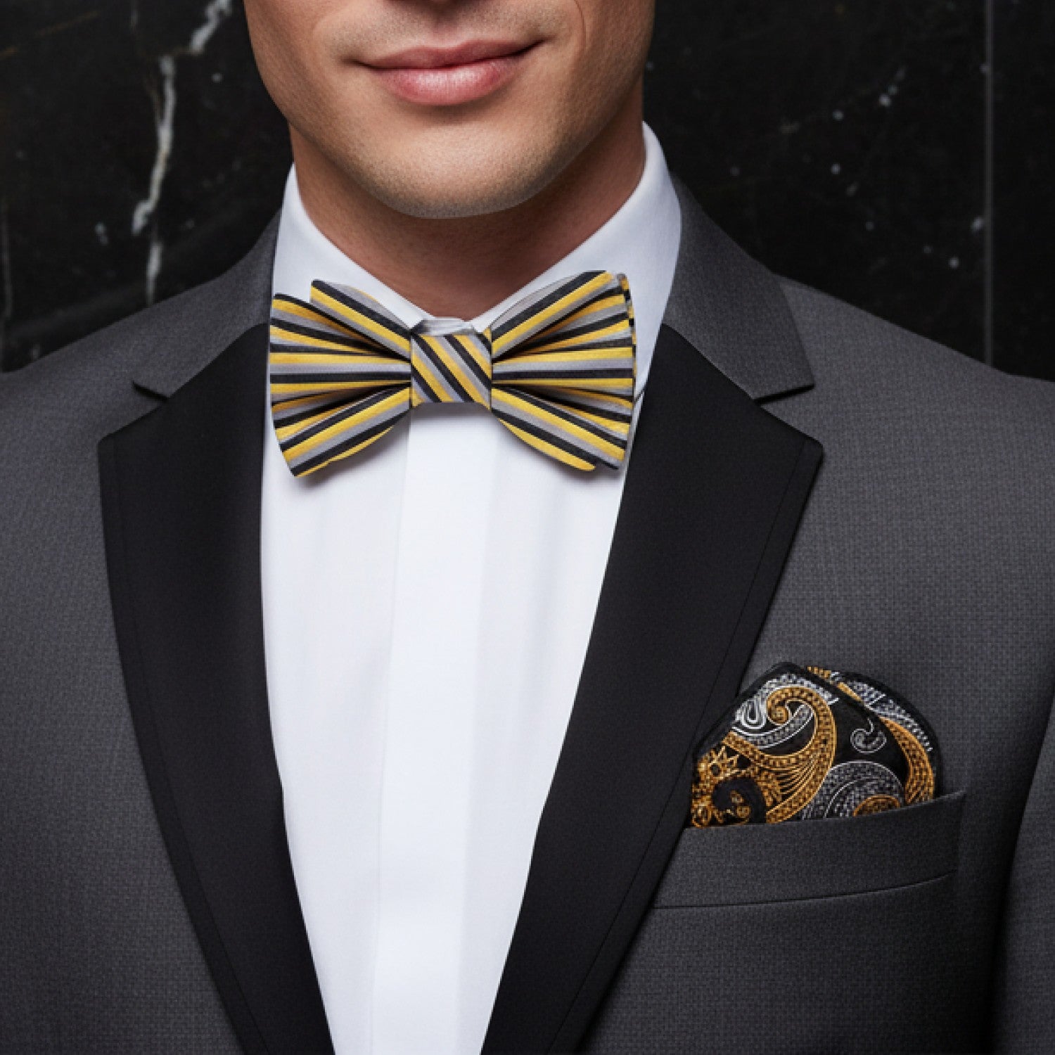Man wearing a gray tuxedo with a yellow striped bow tie and pocket square against a dark background.