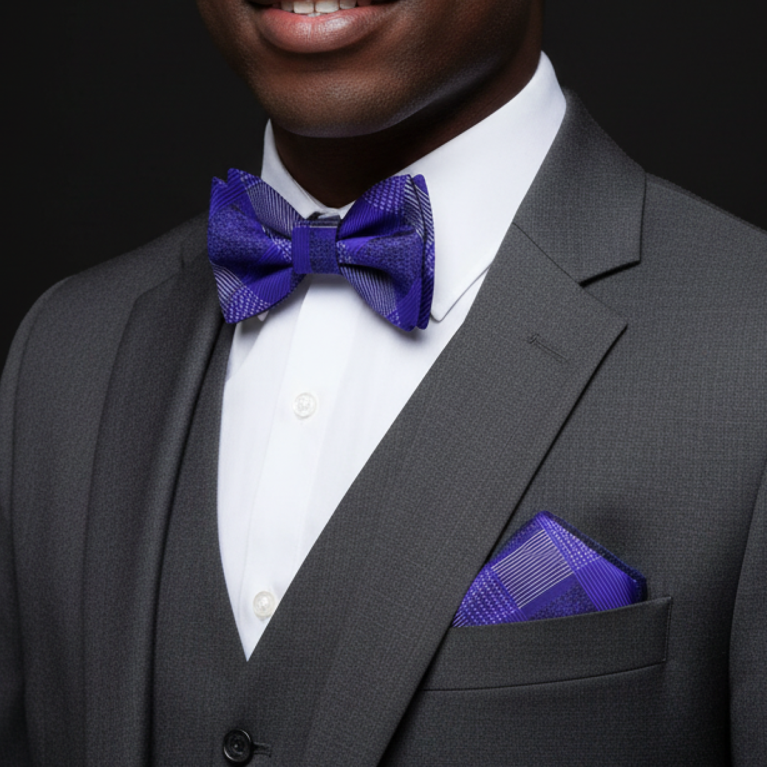 Man wearing a gray suit with a white shirt, purple bow tie, and matching pocket square against a black background.