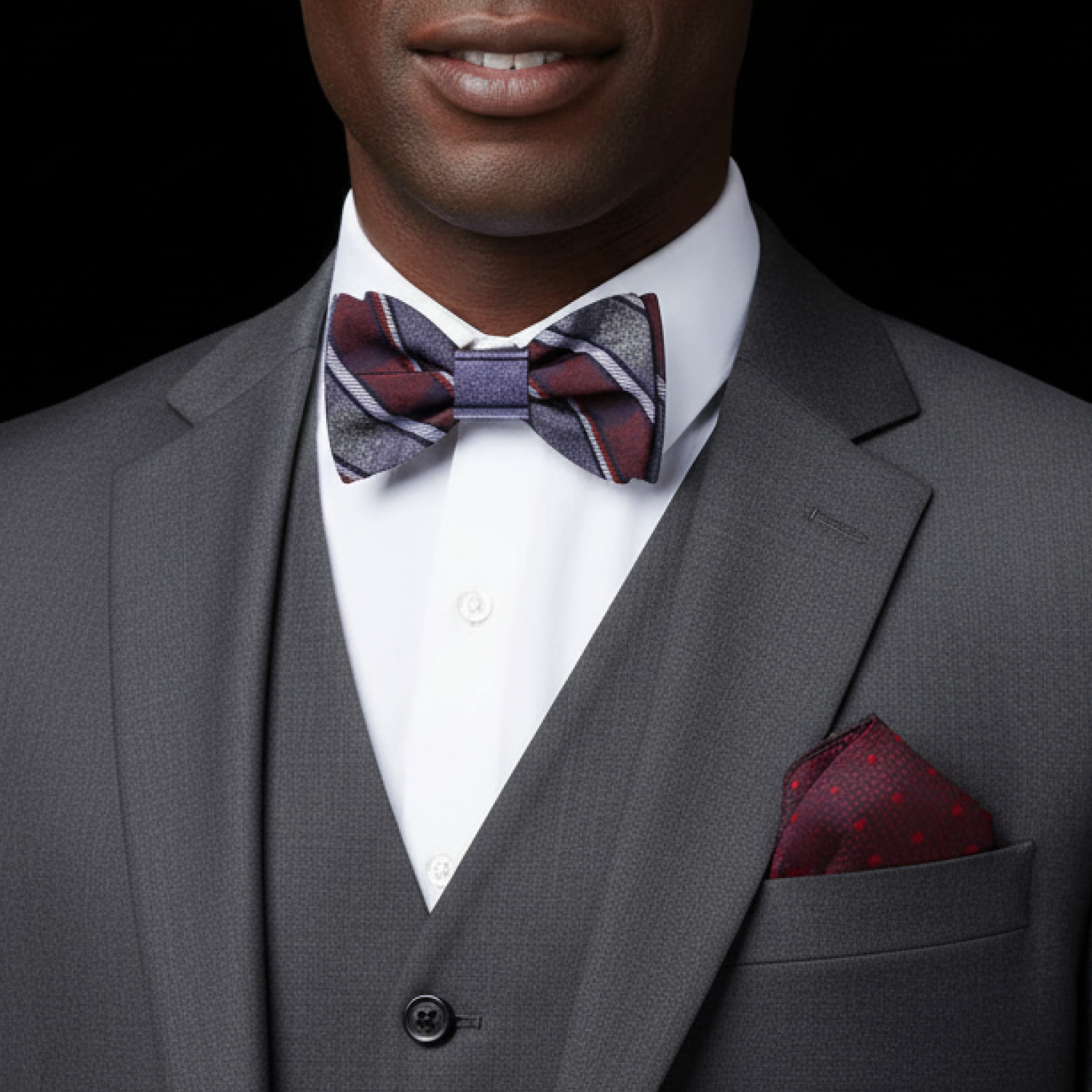 Man wearing a gray suit with a red and grey stripe plaid patterned bow tie and accenting pocket square on a black background