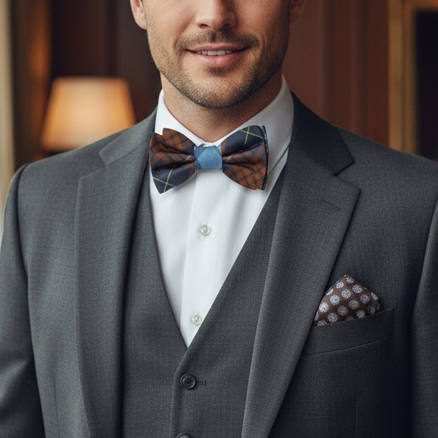 Man wearing a gray suit with a patterned bow tie and pocket square indoors.